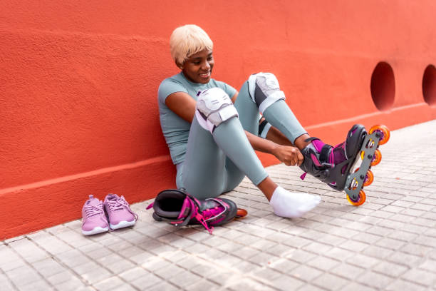 Smiling blonde woman fastening inline skates and knee pads against a red wall.